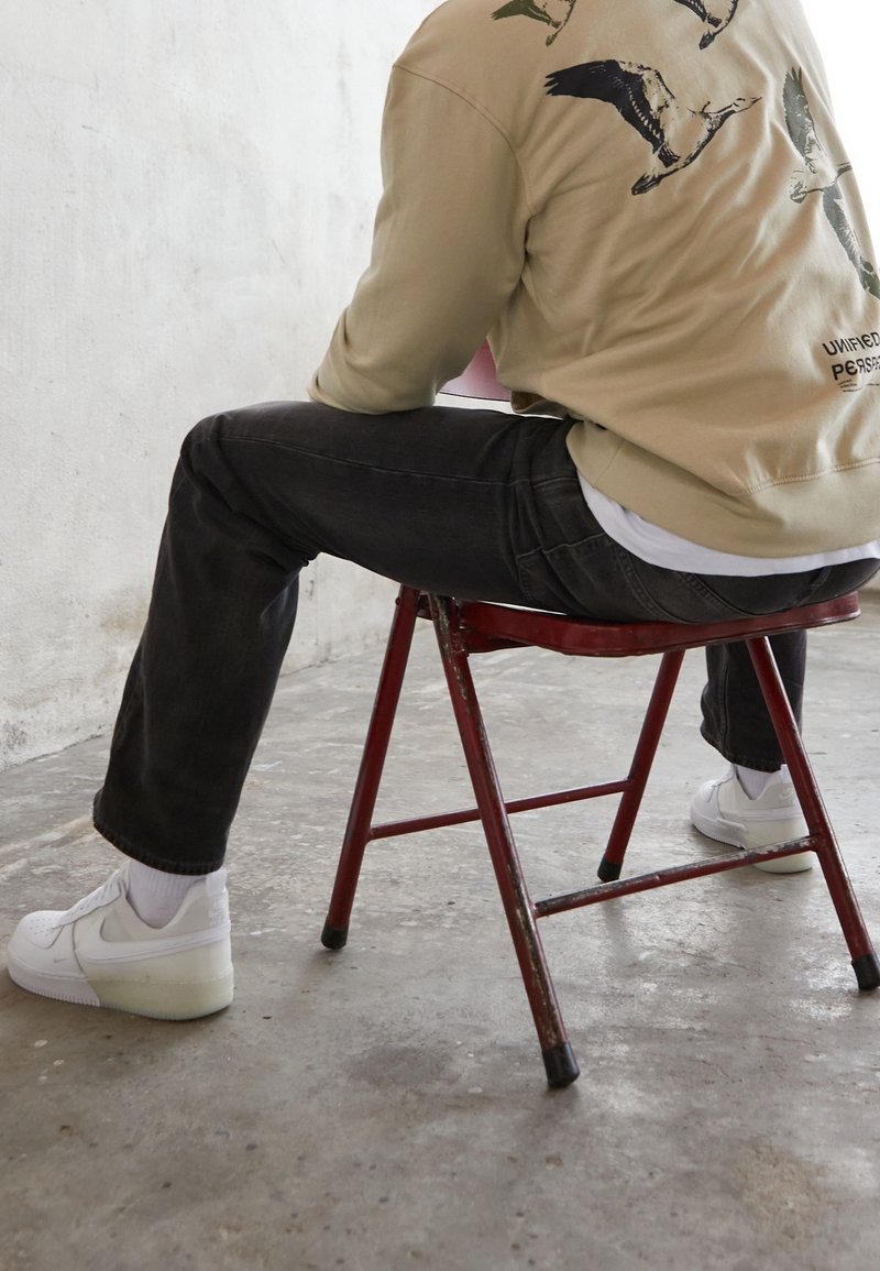 Personne portant un sweat-shirt beige avec des imprimés d'oiseaux, un jean noir et des baskets blanches, assise sur une chaise pliante en métal rouge sur un sol en béton.