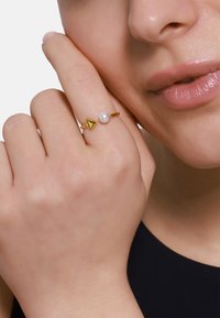 Anello d'oro con design triangolare e una singola perla bianca. La mano è posizionata vicino al viso, mettendo in risalto i suoi delicati tratti.