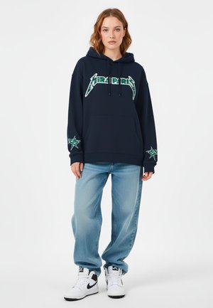Felpa blu navy con grafica frontale e dettagli a stella sulle maniche, abbinata a jeans baggy azzurri e sneakers bianco e nere.