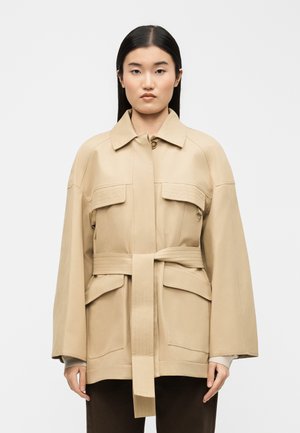 Femme aux cheveux noirs et raides portant une veste beige ceinturée avec de grandes poches avant et un col, debout devant un fond blanc uni.