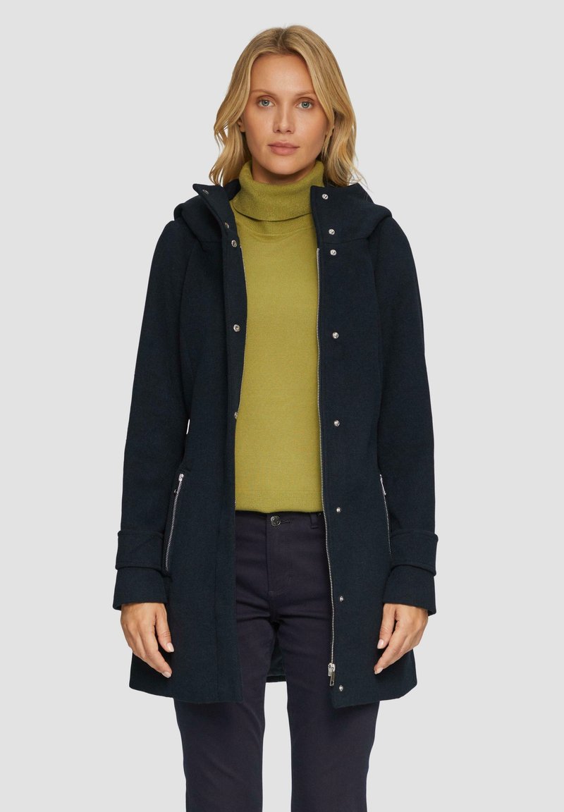 Dunkelblauer Mantel mit Kapuze, ausgestattet mit silbernen Druckknöpfen, seitlichen Reißverschlusstaschen und einer weichen Textur. Getragen über einem grünen Rollkragenpullover.