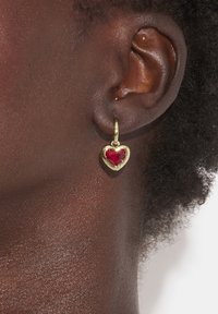 Guldörhänge i hjärtform med en röd ädelsten i mitten. Örhänget är fäst vid en ring och är placerat mot mörk hud och lockigt hår.