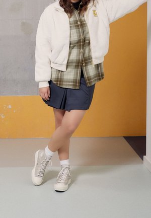 Person iført en off-white fleecejakke, grønternet skjorte, navy nederdel, hvide sokker og grå høje sneakers står indendørs.