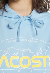 Ljusblå hoodie i bomull, med dragsko i huvan, grafisk kartdesign och texten "LACOSTE INTERNATIONELL" i gult.