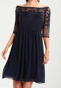 Marinblå klänning med genomskinliga trekvartsärmar, broderad blommönster på bysten, figursydd midja och flödande kjol. Texturerad tyg.