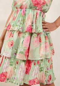Abito floreale con base verde, caratterizzato da strati di volant e motivi di rose rosa. Tessuto leggero e trasparente con una texture plissettata.