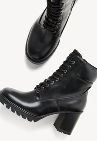 Stivali alla caviglia in pelle nera caratterizzati da un design con lacci, tacco spesso e suola robusta con una finitura texturizzata. Superficie liscia e lucida.