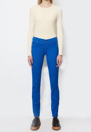 Maglietta a maniche lunghe a coste color crema abbinata a pantaloni aderenti blu. I pantaloni hanno tasche frontali e chiusura con bottone anteriore. Scarpe nere con suola spessa.