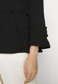 Schwarzes, texturiertes Oberteil mit Rüscheärmel, das ausgefranste Kanten aufweist; kombiniert mit hellen Hosen, wobei eine Hand am unteren Bildrand sichtbar ist.