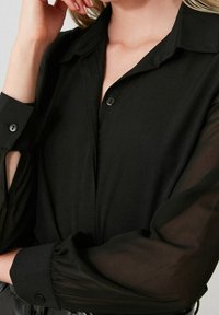 Camisetta nera con colletto, maniche lunghe trasparenti e polsini abbottonati, caratterizzata da una texture di tessuto liscia e un design minimalista.