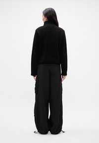 Svart, kort fleecejakke med høy krage. Kombinert med løst sittende svarte bukser med sidelommer og teksturerte detaljer.