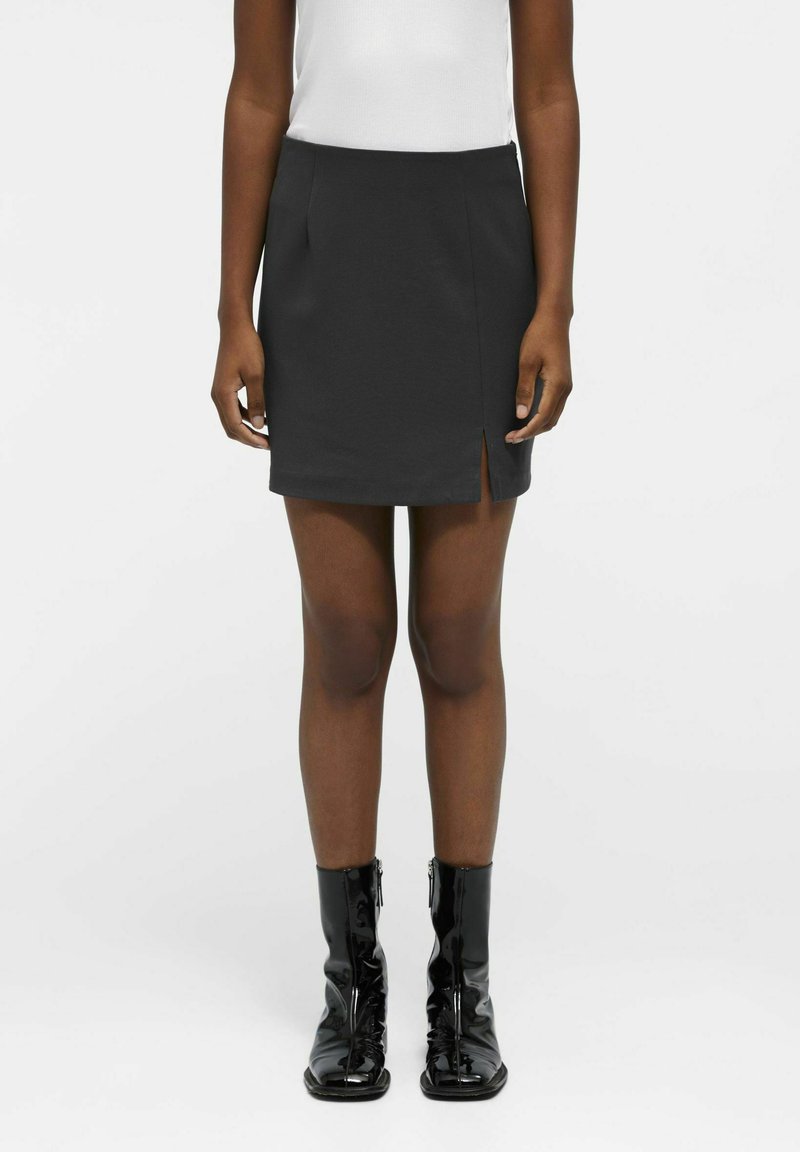 Mini-jupe noire ajustée avec un détail fendu à l'avant. Tissu lisse, design minimaliste. Accompagnée de bottines noires brillantes avec des zips.