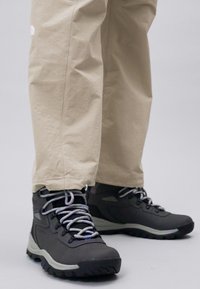 Black hiking boots with gray accents and laces, featuring a rubber sole and a high-top design, paired with beige, loose-fitting pants.