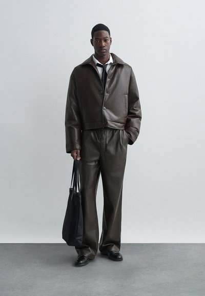 Jachetă din piele maro, scurtă, cu închidere cu nasture, asortată cu pantaloni din piele maro cu șnur. O geantă mare neagră și pantofi completează aspectul.