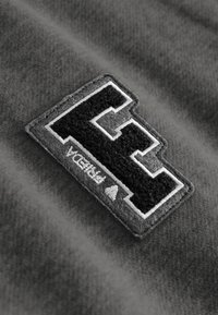 Zwarte fleece patch met een gestileerde letter "E" omlijnd met witte draad, gestikt op een grijze stoffen achtergrond.