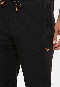 Zwarte sweatpants met een elastische tailleband en zichtbare oranje trekkoord. Heeft zijzakken en een klein oranje logo op de bovenbeen.
