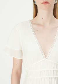 Vestido branco com um decote em V profundo e mangas levemente ribeteadas, acentuado com rendas intrincadas e um design de cintura franzida.
