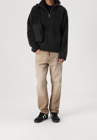 Sudadera de felpa negra con media cremallera, combinada con jeans beige claros y zapatillas negras. Una pequeña bolso negro de hombro cuelga del lado izquierdo.