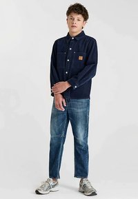 Marineblauw denim overhemd met knoopsluiting, twee borstzakken en een merkpatch, gecombineerd met lichtblauwe distressed jeans en grijze sportschoenen.