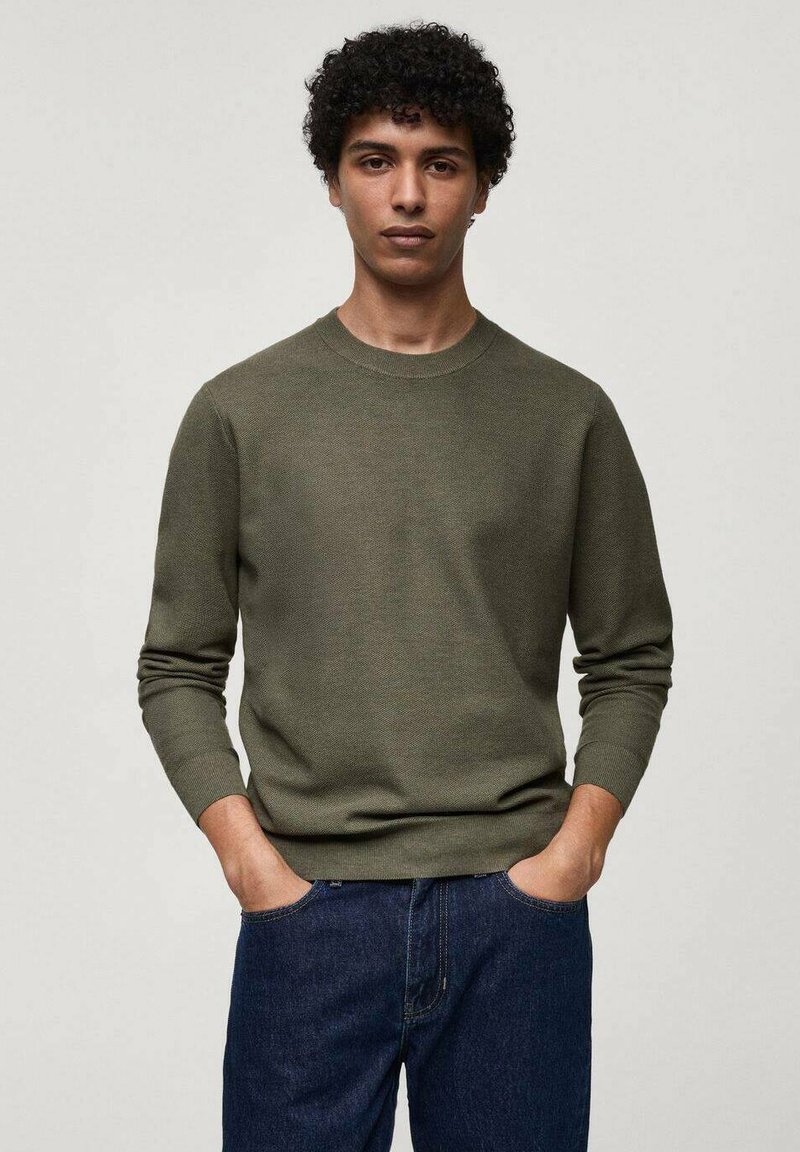 Pullover in maglia verde oliva con scollatura a girocollo, maniche lunghe, polsini a coste e finitura testurizzata, abbinato a jeans in denim scuro.