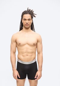 Μυώδης άνδρας με μακριά dreadlocks στέκεται στραμμένος προς τα εμπρός φορώντας μαύρα μπόξερ Calvin Klein μπροστά σε απλό λευκό φόντο.