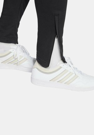 Joggers negros con cremalleras laterales, combinados con zapatillas blancas que tienen tres franjas beiges y suelas texturizadas. Calcetines visibles sobre los zapatos.