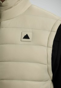 Gilet trapuntato beige con una toppa a forma di montagna nera sul petto, indossato sopra un indumento a maniche lunghe nera.