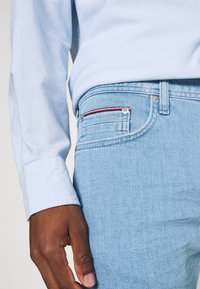 Camisa de manga larga azul claro combinada con jeans azul claro que presentan un acento rojo en el bolsillo, mostrando la textura y el ajuste del denim.