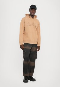 Beige hoodies med framfickor och grafisk design, i kombination med svarta cargobyxor med ett urblekt mönster och flera fickor. Svarta sneakers.