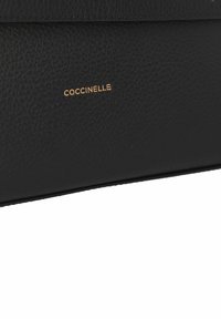 Borsa a tracolla in pelle nera con texture e logo "Coccinelle" in rilievo dorato. Il design presenta linee pulite e un'estetica minimalista.