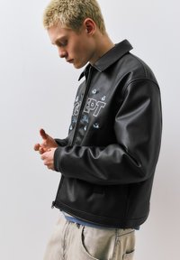 Mladá osoba s krátkými blond vlasy, oblečená v černé kožené bundě s nápisem "CONCEPT" a světle zbarvených kalhotách, dívající se dolů na své ruce.