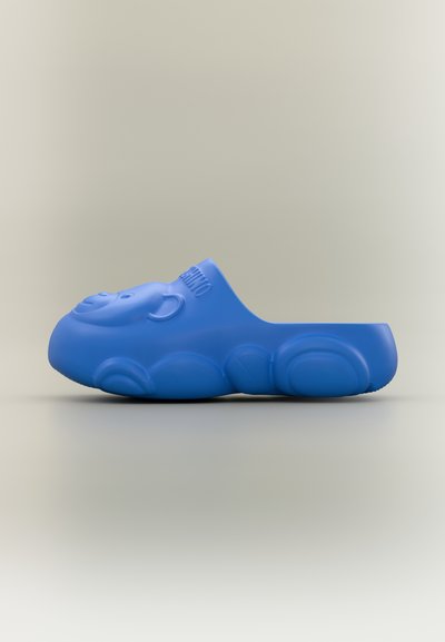 Sandale bleu à enfiler en forme de tête d'ours souriante avec des contours arrondis et un texte embossé sur la partie supérieure.