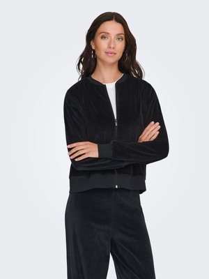 Femme aux longs cheveux foncés portant des boucles d'oreilles créoles argentées, une veste noire zippée et un pantalon noir assorti, bras croisés, fond neutre.