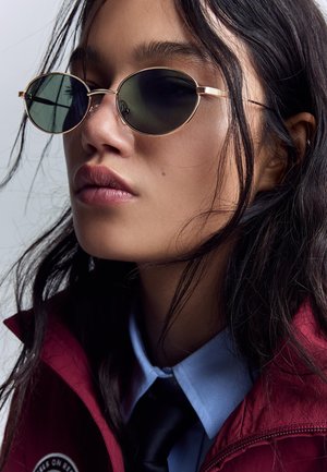 Giovane donna con lunghi capelli scuri che indossa occhiali da sole ovali, una giacca rossa, una camicia blu con colletto e una cravatta nera, che guarda leggermente di lato.