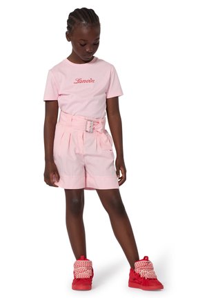 Bambino che indossa pantaloncini rosa con cintura, maglietta rosa con scritta rossa "Lanvin" e scarpe da ginnastica alte rosse con lacci bianchi con motivi, in piedi con la testa abbassata.