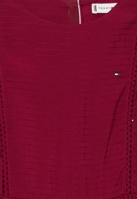 Bordová blúzka s textúrovaným vertikálnym pruhovaním, so čipkovými detailmi po stranách a malým logom na hrudi.