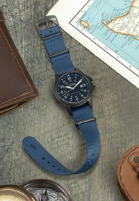 Blå nylon rem ur med en sort plasturkasse og en blå skive. Har store hvide tal, dato display og sølvfarvede accenter.