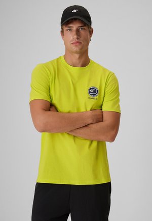 Mężczyzna ze skrzyżowanymi ramionami, ubrany w jasny żółty t-shirt z okrągłym logo i czarną czapką, stojący na tle jednolitego szarego tła.