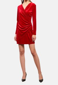 Robe en velours rouge avec des manches longues et un décolleté en V. Présente un tissu drapé et une silhouette ajustée, se terminant au-dessus du genou. Escarpins noirs.