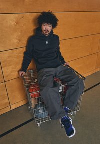 Svart hettegenser med logo, grå cargo-bukser og blå sneakers. Sittende i en kurv av metall med basketballer på gymgulvet.
