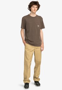 Brown cotton t-shirt with a chest pocket. Paired with beige trousers and black sneakers featuring a mesh design. Positioned in a frontal stance.