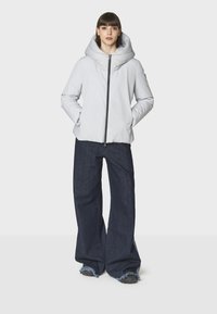 Giacca puffer grigia chiara con un grande cappuccio, chiusura con zip, tasche laterali e pantaloni ampi in denim scuro con orli sfilacciati.