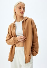 Sudadera marrón con cremallera y cordones, de tejido suave, corte relajado, combinada con una blusa corta blanca y pantalones de color claro.