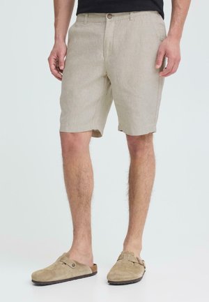 Mand iført beige knælange shorts og matchende slip-on ruskindssko med sorte såler, stående mod en ensfarvet baggrund.