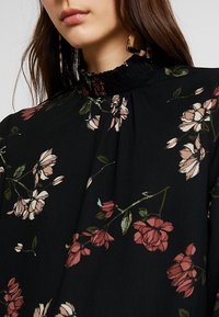 Schwarze Bluse mit Blumenmuster, die einen hohen gerafften Kragen hat, mit rosa und cremefarbenen Blumen sowie grünen Blättern. Leichter Stoff mit strukturierter Oberfläche.