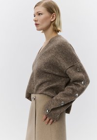 Pulover maro tricotat cu decolteu în V și manșete cu model nervurat, cu nasturi argintii pe mâneci, asortat cu o fustă bej.
