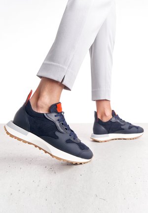 Persona che indossa sneakers blu navy e bianche con dettagli arancioni e pantaloni grigio chiaro, mentre calpesta una superficie grigio chiaro.