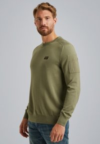 Olivno zeleno bombažno pulover s rebrastim ovratnikom in manšeti, ki ima majhno nalepko na prsih in teksturirane ramenske poudarke. Preprost, prilegajoč se dizajn.