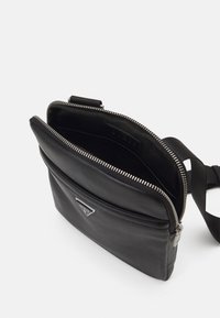 Borsa a tracolla in pelle nera con chiusura a zip, dotata di logo triangolare e tasca frontale, texture liscia e tracolla regolabile.