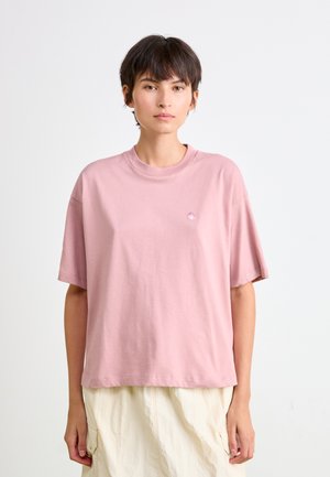 Young person with short dark hair wearing a loose pink t-shirt and cream cargo-style pants against a plain white background.